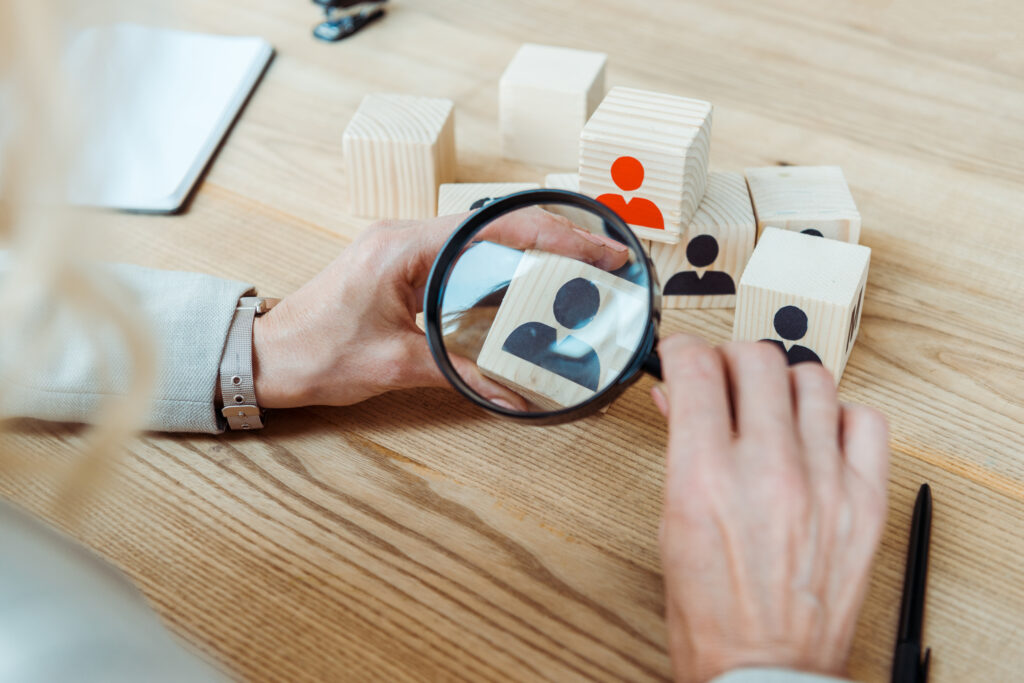 Recruiter gebruikt een vergrootglas om een houten blokje met een icoon van een persoon te inspecteren, symbool voor het vinden van de juiste match.