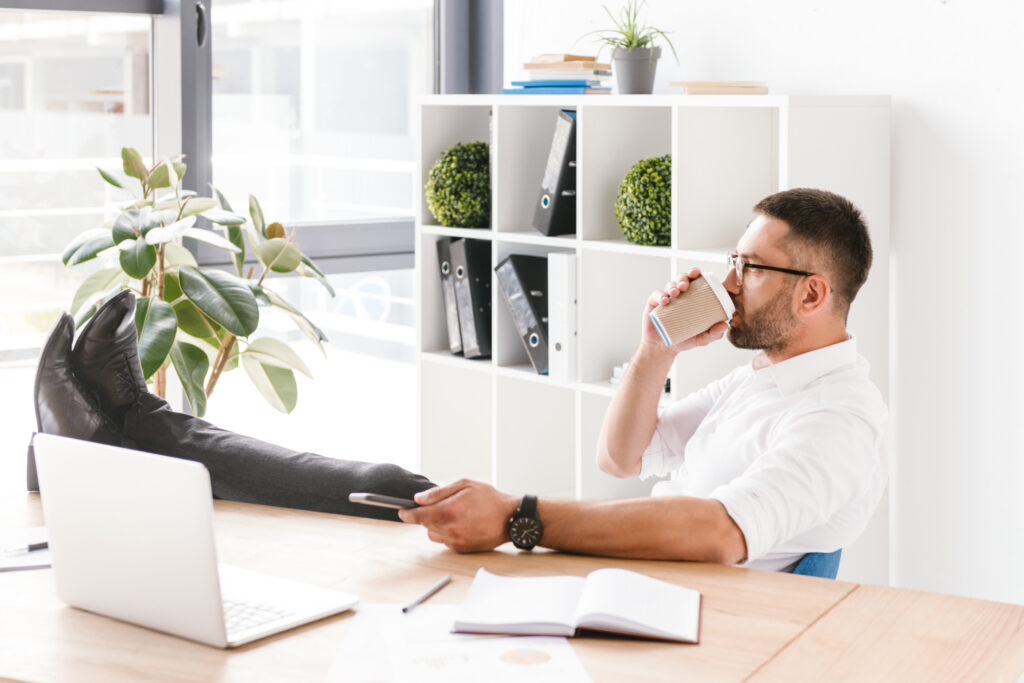 Man in formele kleding drinkt koffie terwijl hij ontspannen werkt in een modern kantoor, met benen op tafel.